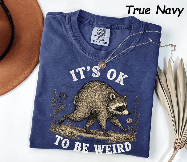 May include: A true navy blue short-sleeve t-shirt featuring a graphic of a raccoon walking through a field with mushrooms and flowers. The text "IT'S OK TO BE WEIRD" is printed in white above and below the raccoon. The shirt has a Comfort Colors label.