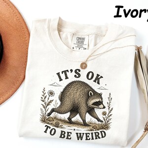 May include: An ivory t-shirt featuring a graphic illustration of a raccoon walking through a field of flowers and mushrooms. The text "IT'S OK TO BE WEIRD" is printed in black lettering above and below the raccoon. The shirt has a Comfort Colors label.