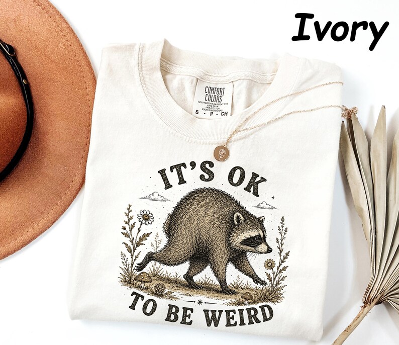 May include: An ivory t-shirt featuring a graphic illustration of a raccoon walking through a field of flowers and mushrooms. The text "IT'S OK TO BE WEIRD" is printed in black lettering above and below the raccoon. The shirt has a Comfort Colors label.