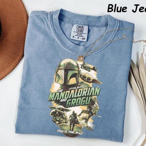 May include: Blue jean colored t-shirt featuring a graphic of The Mandalorian and Grogu from Star Wars. The shirt has a crew neck and short sleeves. A gold necklace with a pendant is draped across the shirt. The text "THIS IS THE WAY" is printed on the shirt.
