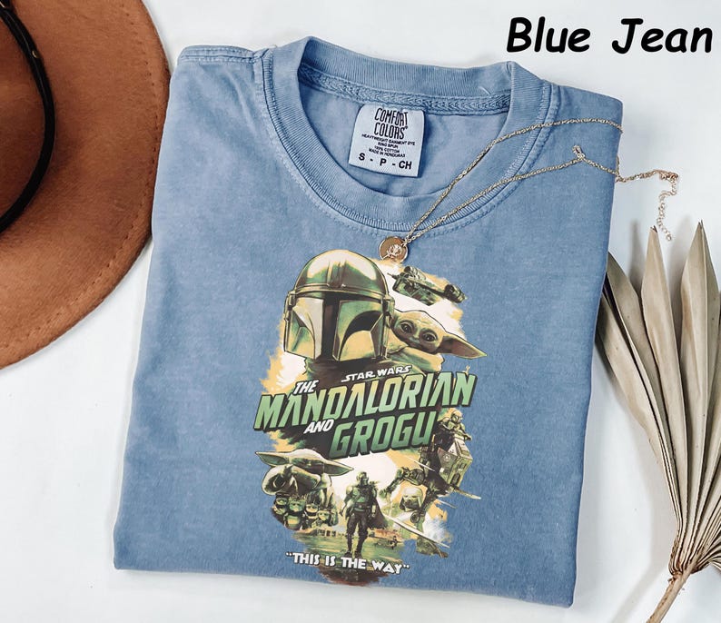 May include: Blue jean colored t-shirt featuring a graphic of The Mandalorian and Grogu from Star Wars. The shirt has a crew neck and short sleeves. A gold necklace with a pendant is draped across the shirt. The text "THIS IS THE WAY" is printed on the shirt.