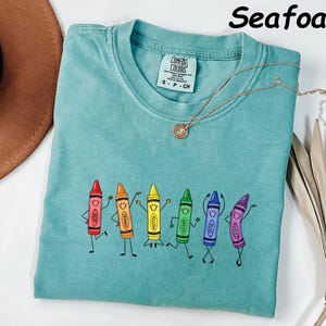 May include: A seafoam green short-sleeve t-shirt featuring a graphic of six anthropomorphic crayons in red, orange, yellow, green, blue, and violet, each with a smiling face and limbs. The shirt has a crew neckline and a Comfort Colors brand label.