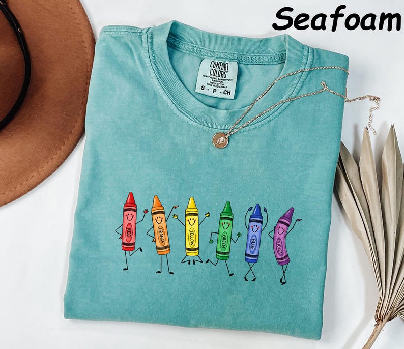 May include: A seafoam green short-sleeve t-shirt featuring a graphic of six anthropomorphic crayons in red, orange, yellow, green, blue, and violet, each with a smiling face and limbs. The shirt has a crew neckline and a Comfort Colors brand label.