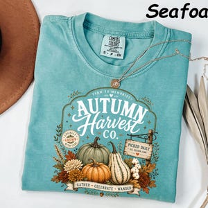 May include: A seafoam green short-sleeve t-shirt featuring a graphic print with the text Autumn Harvest Co. Farm to Memories, Locally Grown with Care, and Picked Daily All Season Long. The design includes pumpkins, gourds, and autumn foliage.