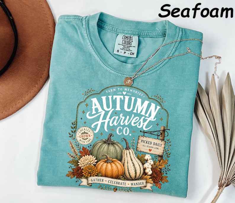 May include: A seafoam green short-sleeve t-shirt featuring a graphic print with the text Autumn Harvest Co. Farm to Memories, Locally Grown with Care, and Picked Daily All Season Long. The design includes pumpkins, gourds, and autumn foliage.