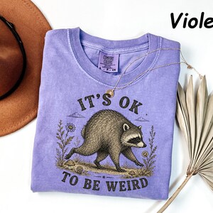 May include: A violet short-sleeved t-shirt featuring a graphic illustration of a raccoon walking among flowers and mushrooms. The text "IT'S OK TO BE WEIRD" is printed in black above and below the raccoon. The shirt has a Comfort Colors label.