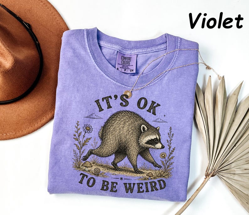 May include: A violet short-sleeved t-shirt featuring a graphic illustration of a raccoon walking among flowers and mushrooms. The text "IT'S OK TO BE WEIRD" is printed in black above and below the raccoon. The shirt has a Comfort Colors label.