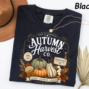 May include: A black t-shirt featuring a graphic with the text "Autumn Harvest Co." and "Farm to Memories." The design includes illustrations of pumpkins, gourds, autumn leaves, and flowers, along with a sign reading "Picked Daily."