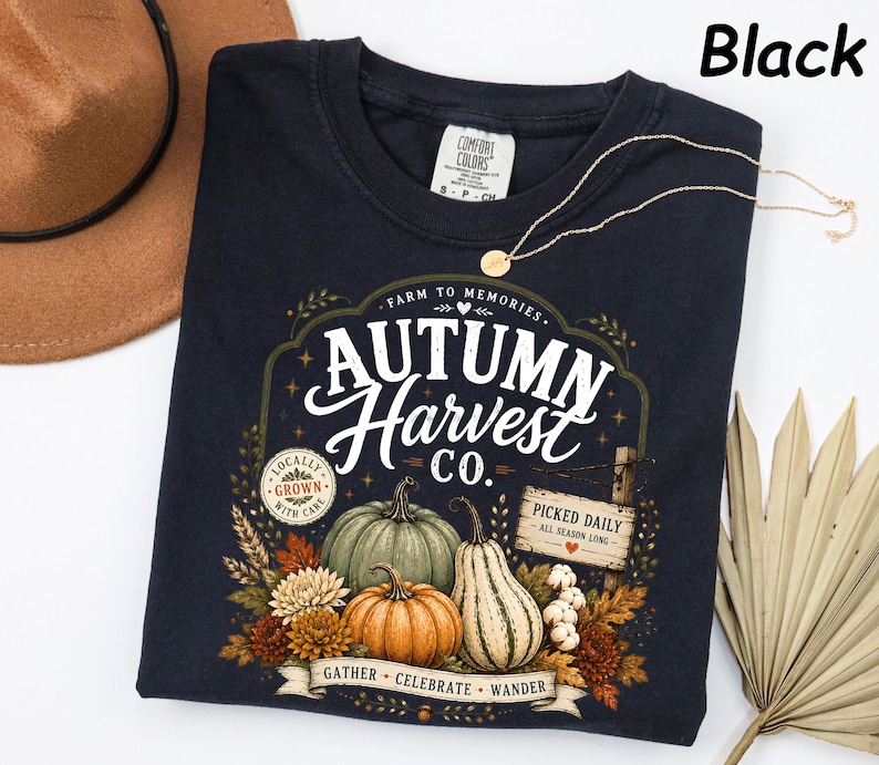 May include: A black t-shirt featuring a graphic with the text "Autumn Harvest Co." and "Farm to Memories." The design includes illustrations of pumpkins, gourds, autumn leaves, and flowers, along with a sign reading "Picked Daily."
