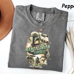 May include: A gray t-shirt featuring a graphic from the Star Wars series, "The Mandalorian and Grogu." The design includes the Mandalorian's helmet and Grogu. The text "THIS IS THE WAY" is at the bottom. A silver necklace with a pendant is on the shirt.