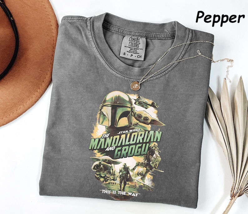 May include: A gray t-shirt featuring a graphic from the Star Wars series, "The Mandalorian and Grogu." The design includes the Mandalorian's helmet and Grogu. The text "THIS IS THE WAY" is at the bottom. A silver necklace with a pendant is on the shirt.