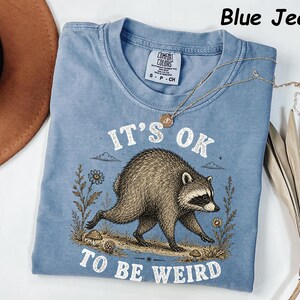 May include: A blue jean coloured t-shirt featuring a graphic of a raccoon walking through a field with flowers and mushrooms. The text above the raccoon reads IT'S OK and the text below reads TO BE WEIRD. The shirt has a Comfort Colors brand label.