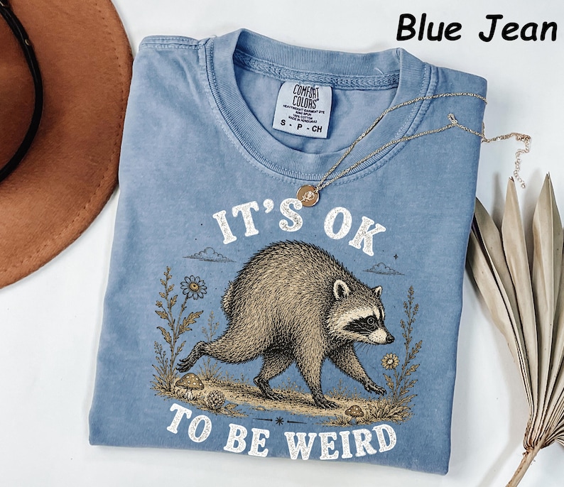 May include: A blue jean coloured t-shirt featuring a graphic of a raccoon walking through a field with flowers and mushrooms. The text above the raccoon reads IT'S OK and the text below reads TO BE WEIRD. The shirt has a Comfort Colors brand label.