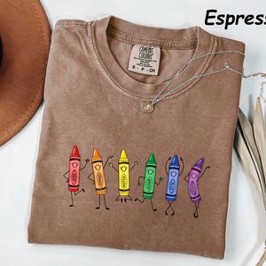 May include: An espresso-colored t-shirt featuring a graphic of six anthropomorphic crayons in red, orange, yellow, green, blue, and violet. Each crayon has a smiling face and stick-figure limbs. The shirt has a crew neck and a Comfort Colors label.