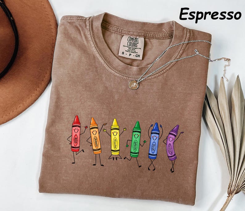 May include: An espresso-colored t-shirt featuring a graphic of six anthropomorphic crayons in red, orange, yellow, green, blue, and violet. Each crayon has a smiling face and stick-figure limbs. The shirt has a crew neck and a Comfort Colors label.