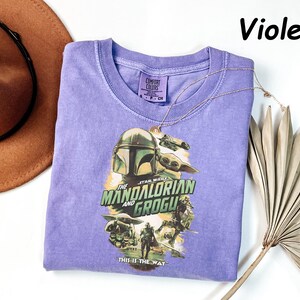 May include: Violet-colored t-shirt featuring a graphic from "The Mandalorian" with the text "The Mandalorian and Grogu" and "This is the Way." The shirt has a crew neck and short sleeves. A gold necklace and a brown hat are also in the image.