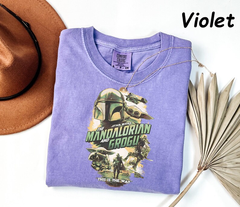 May include: Violet-colored t-shirt featuring a graphic from "The Mandalorian" with the text "The Mandalorian and Grogu" and "This is the Way." The shirt has a crew neck and short sleeves. A gold necklace and a brown hat are also in the image.