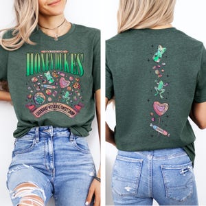 May include: Dark green t-shirt with a graphic of the Honeydukes sweet shop. The front features the text "Honeydukes" and various candies. The back has a vertical design with a gummy bear, frog, and heart-shaped lollipop.