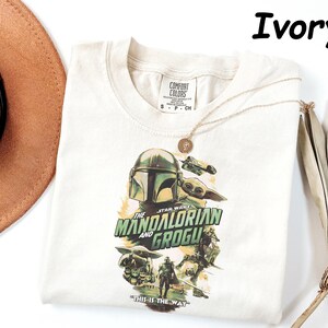 May include: Ivory-colored t-shirt featuring a graphic of The Mandalorian and Grogu from Star Wars. The design includes the Mandalorian's helmet, Grogu, and the text "THE MANDALORIAN AND GROGU" and "THIS IS THE WAY". A gold necklace is draped across the shirt.