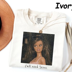 May include: An ivory short-sleeved t-shirt with a crew neckline and a Comfort Colors brand label. The center features a square graphic of a doll with long brown hair and a pink top, with the text "y'all need Jesus." printed below the image.