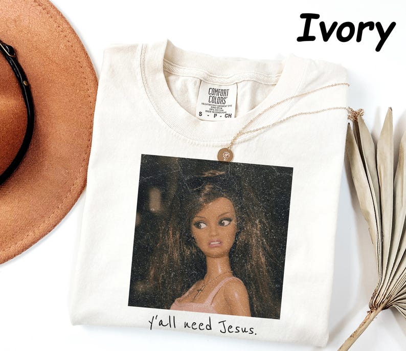 May include: An ivory short-sleeved t-shirt with a crew neckline and a Comfort Colors brand label. The center features a square graphic of a doll with long brown hair and a pink top, with the text "y'all need Jesus." printed below the image.