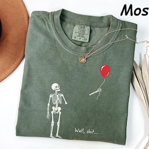 May include: A moss green t-shirt featuring a white graphic of a skeleton looking up at a red balloon held by a detached skeletal hand. The text "Well, shit..." is printed in white below the skeleton. The shirt has a Comfort Colors brand label.
