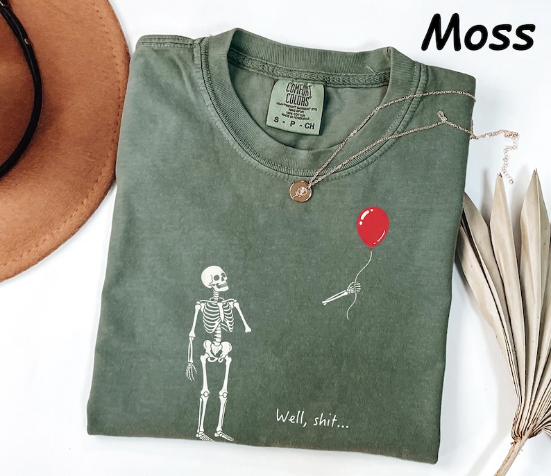 May include: A moss green t-shirt featuring a white graphic of a skeleton looking up at a red balloon held by a detached skeletal hand. The text "Well, shit..." is printed in white below the skeleton. The shirt has a Comfort Colors brand label.