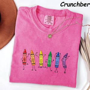 May include: A pink short-sleeved t-shirt featuring a graphic of six anthropomorphic crayons in red, orange, yellow, green, blue, and violet, each with a smiling face and stick limbs. The shirt has a crew neckline and a Comfort Colors brand label.