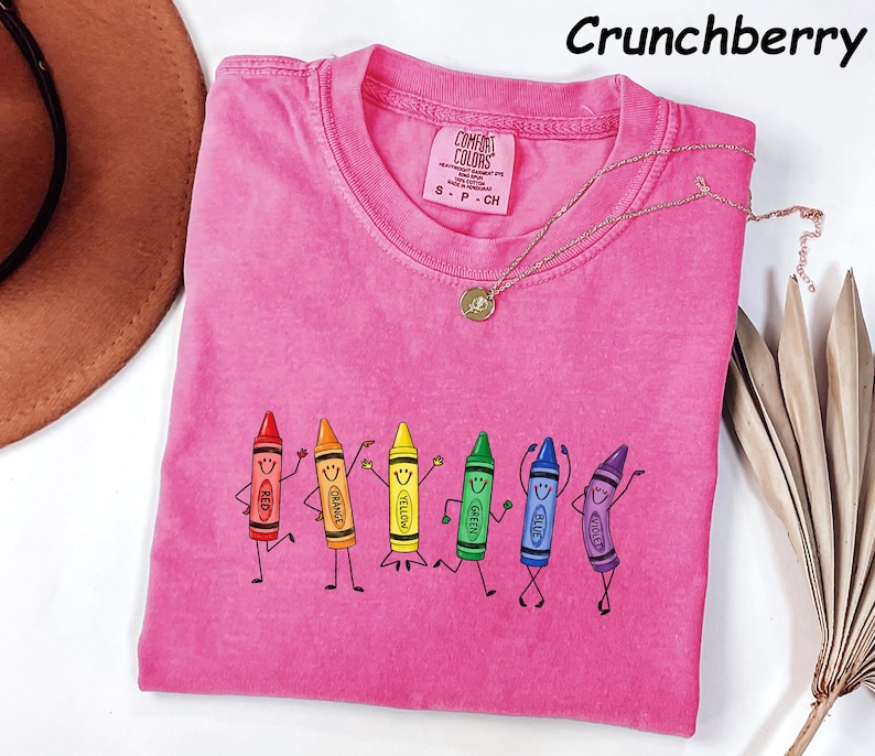 May include: A pink short-sleeved t-shirt featuring a graphic of six anthropomorphic crayons in red, orange, yellow, green, blue, and violet, each with a smiling face and stick limbs. The shirt has a crew neckline and a Comfort Colors brand label.