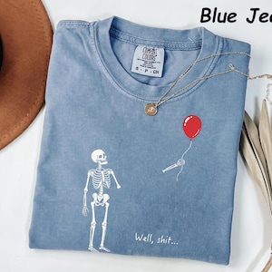 May include: A blue jean colored t-shirt featuring a white graphic of a skeleton looking at a detached skeletal arm holding a red balloon. The text "Well, shit..." is printed in white below the arm. The shirt has a Comfort Colors brand label.