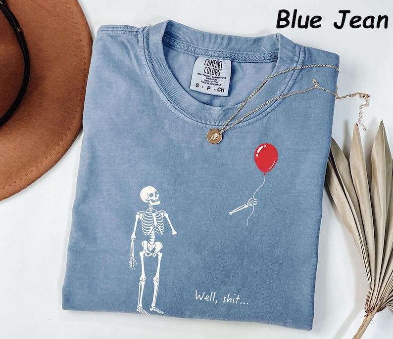 May include: A blue jean colored t-shirt featuring a white graphic of a skeleton looking at a detached skeletal arm holding a red balloon. The text "Well, shit..." is printed in white below the arm. The shirt has a Comfort Colors brand label.