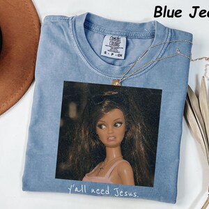 May include: A blue jean colored t-shirt featuring a graphic print of a doll with long brown hair wearing sunglasses and a pink top. The text "y'all need Jesus." is printed below the graphic. The shirt has a Comfort Colors brand label.