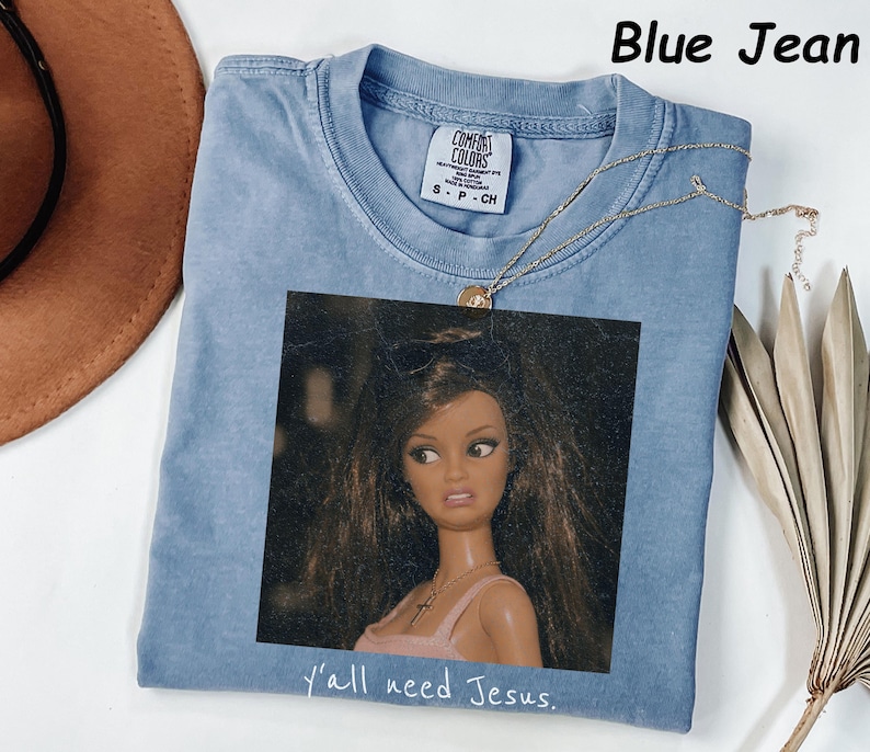 May include: A blue jean colored t-shirt featuring a graphic print of a doll with long brown hair wearing sunglasses and a pink top. The text "y'all need Jesus." is printed below the graphic. The shirt has a Comfort Colors brand label.