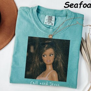 May include: A seafoam green short-sleeve t-shirt featuring a graphic print of a doll with long brown hair wearing sunglasses on her head and a cross necklace. The text "y'all need Jesus." is printed in white below the graphic. The shirt has a Comfort Colors brand label.
