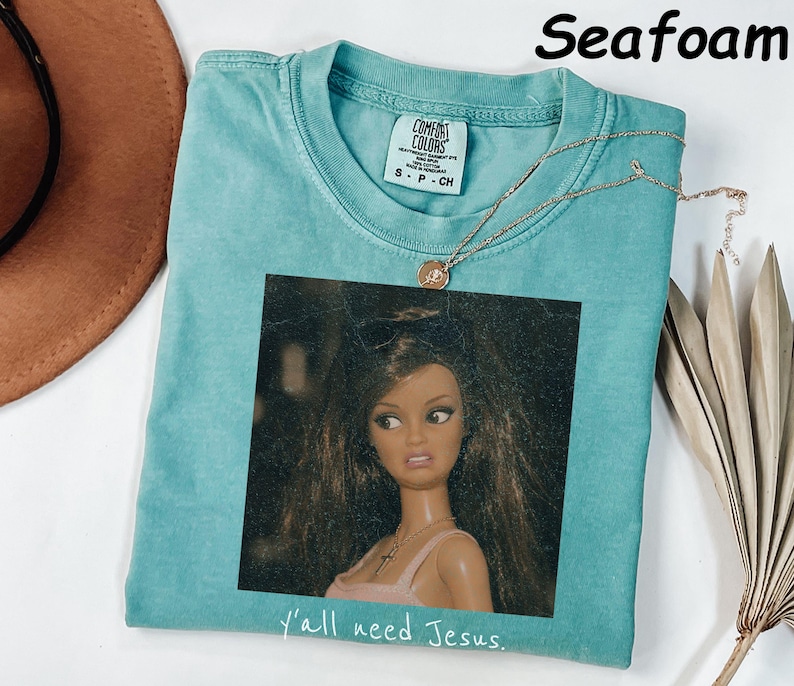 May include: A seafoam green short-sleeve t-shirt featuring a graphic print of a doll with long brown hair wearing sunglasses on her head and a cross necklace. The text "y'all need Jesus." is printed in white below the graphic. The shirt has a Comfort Colors brand label.