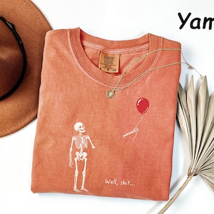 May include: A yam-colored t-shirt featuring a white graphic of a skeleton looking at a floating red balloon. The text "Well, shit..." is printed in white near the bottom. The shirt has a crew neckline and a Comfort Colors brand label.