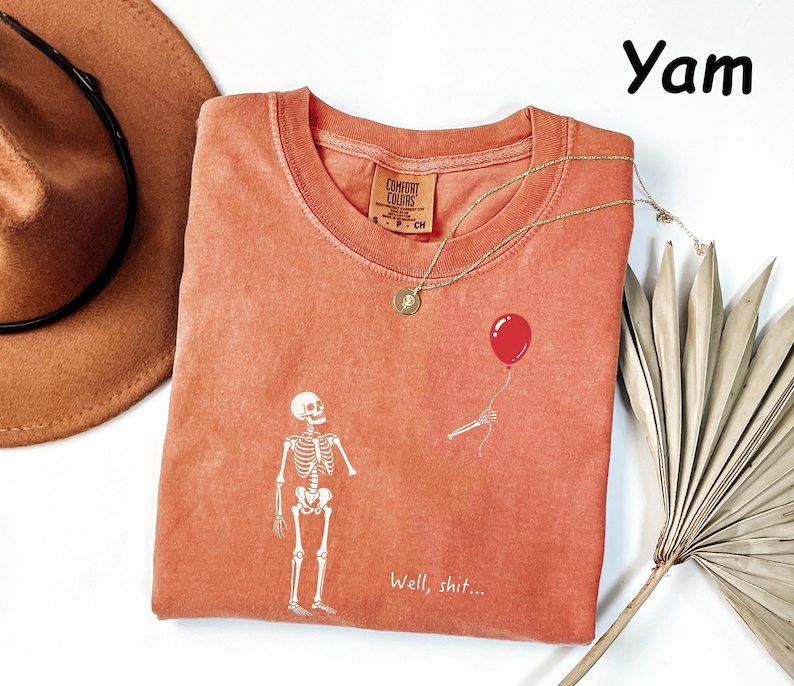 May include: A yam-colored t-shirt featuring a white graphic of a skeleton looking at a floating red balloon. The text "Well, shit..." is printed in white near the bottom. The shirt has a crew neckline and a Comfort Colors brand label.