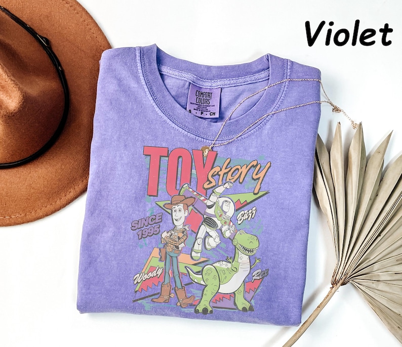 May include: A violet-colored t-shirt featuring a graphic of characters from the movie "Toy Story." The design includes Woody, Buzz Lightyear, and Rex, with the text "TOY STORY" and "SINCE 1995." A gold necklace and a brown hat are also visible.