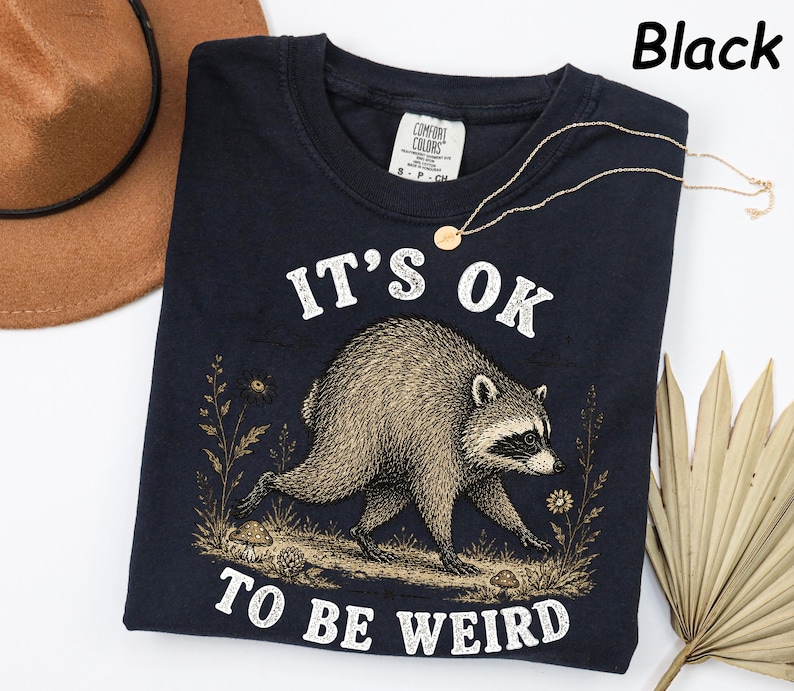 May include: A black t-shirt featuring a graphic illustration of a raccoon walking through a field with mushrooms and flowers. The text "IT'S OK TO BE WEIRD" is printed in white above and below the raccoon. The shirt has a Comfort Colors label.