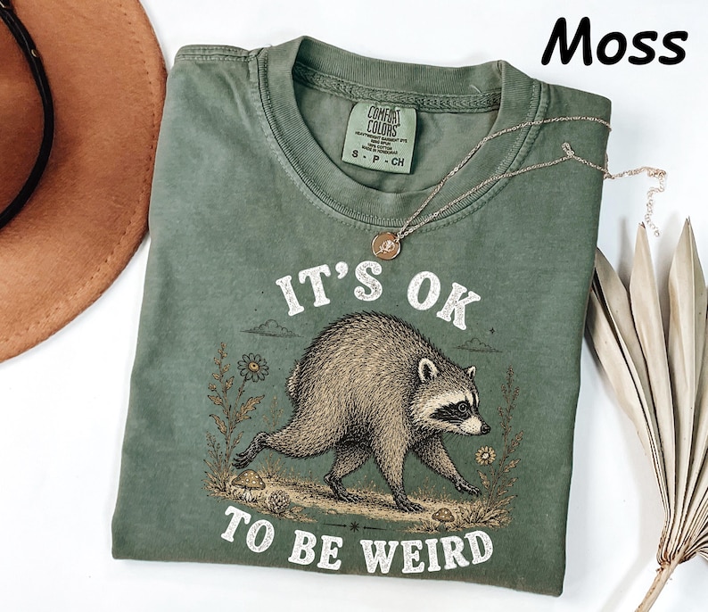 May include: A moss green short-sleeve t-shirt featuring a graphic of a raccoon walking among flowers and mushrooms. The text "IT'S OK TO BE WEIRD" is printed in white above and below the raccoon. The shirt has a Comfort Colors brand label.