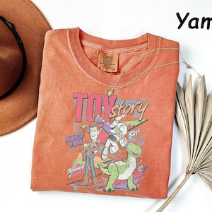 May include: A yam-colored, crew neck t-shirt featuring a graphic of characters from the Toy Story movie. The graphic includes Woody, Buzz Lightyear, and Rex, with the text "TOY STORY" and "SINCE 1995". A gold necklace and a brown hat are also visible.