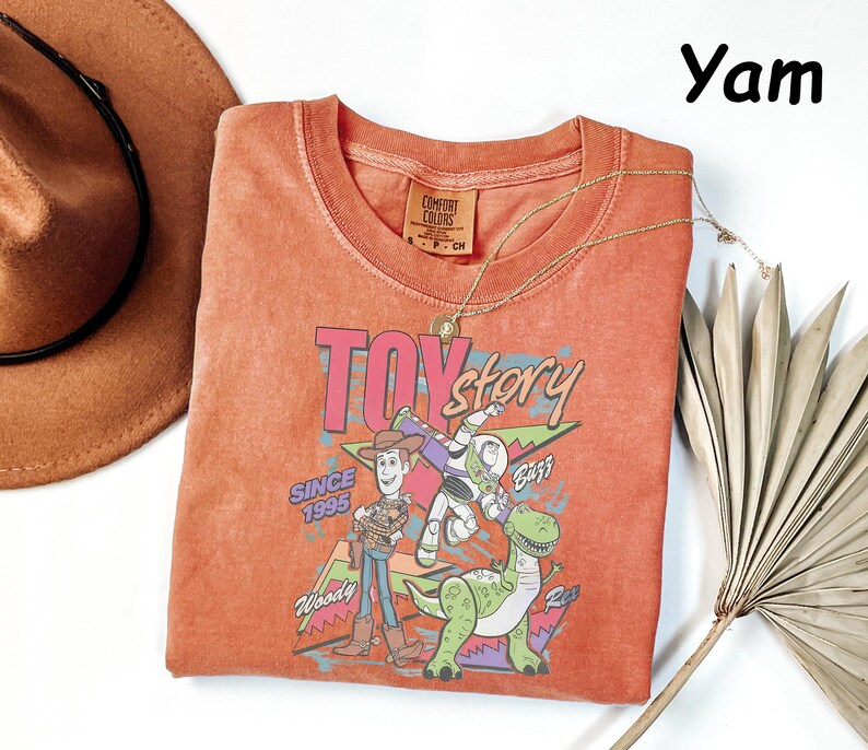 May include: A yam-colored, crew neck t-shirt featuring a graphic of characters from the Toy Story movie. The graphic includes Woody, Buzz Lightyear, and Rex, with the text "TOY STORY" and "SINCE 1995". A gold necklace and a brown hat are also visible.