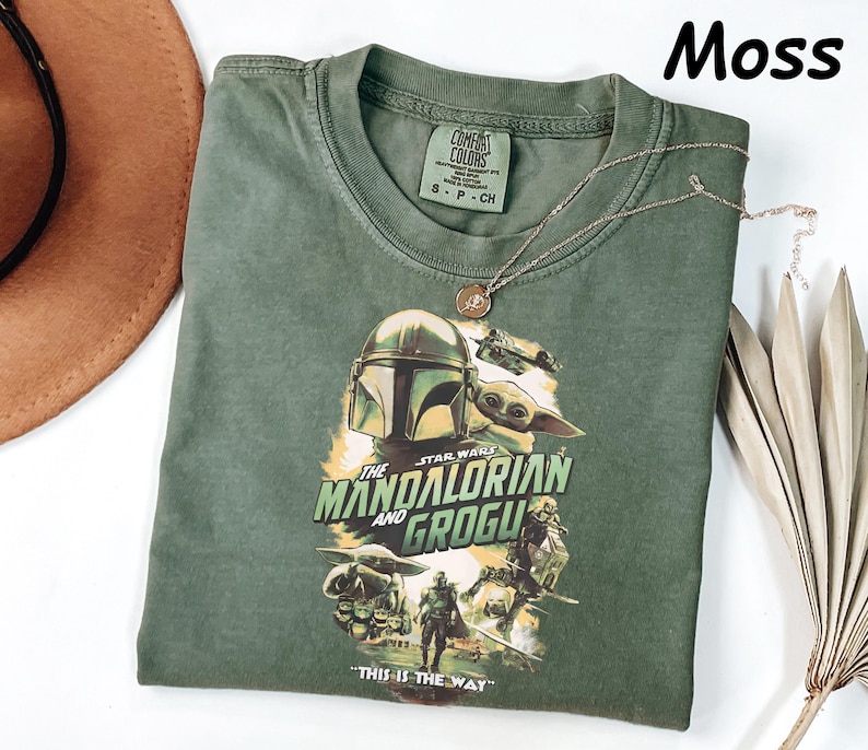May include: A moss green t-shirt featuring a graphic from "The Mandalorian" with the text "The Mandalorian and Grogu" and "This is the Way." A gold-colored necklace is draped across the shirt. The shirt is folded on a white surface.