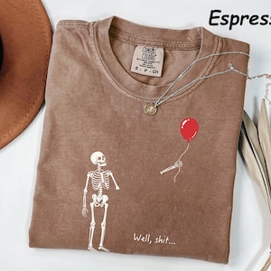 May include: An espresso-colored t-shirt featuring a white graphic of a skeleton looking at a detached skeletal hand holding a red balloon. The text "Well, shit..." is printed in white below the graphic. The shirt has a Comfort Colors brand label.