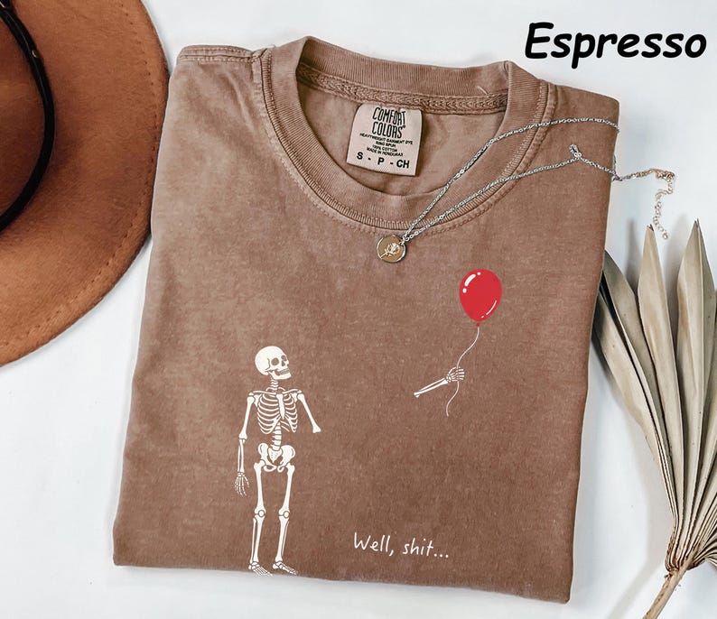 May include: An espresso-colored t-shirt featuring a white graphic of a skeleton looking at a detached skeletal hand holding a red balloon. The text "Well, shit..." is printed in white below the graphic. The shirt has a Comfort Colors brand label.