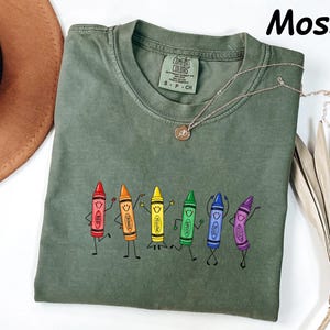 May include: A moss green short-sleeve t-shirt featuring a graphic of six anthropomorphic crayons in red, orange, yellow, green, blue, and violet, each with a smiling face and limbs. The shirt has a crew neckline and a Comfort Colors brand label.