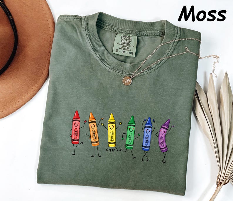 May include: A moss green short-sleeve t-shirt featuring a graphic of six anthropomorphic crayons in red, orange, yellow, green, blue, and violet, each with a smiling face and limbs. The shirt has a crew neckline and a Comfort Colors brand label.