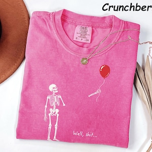 May include: A pink short-sleeved t-shirt featuring a white graphic of a skeleton looking up at a red balloon held by a skeletal hand. The text "Well, shit..." is printed in white below the skeleton. The shirt has a crew neck and a Comfort Colors label.