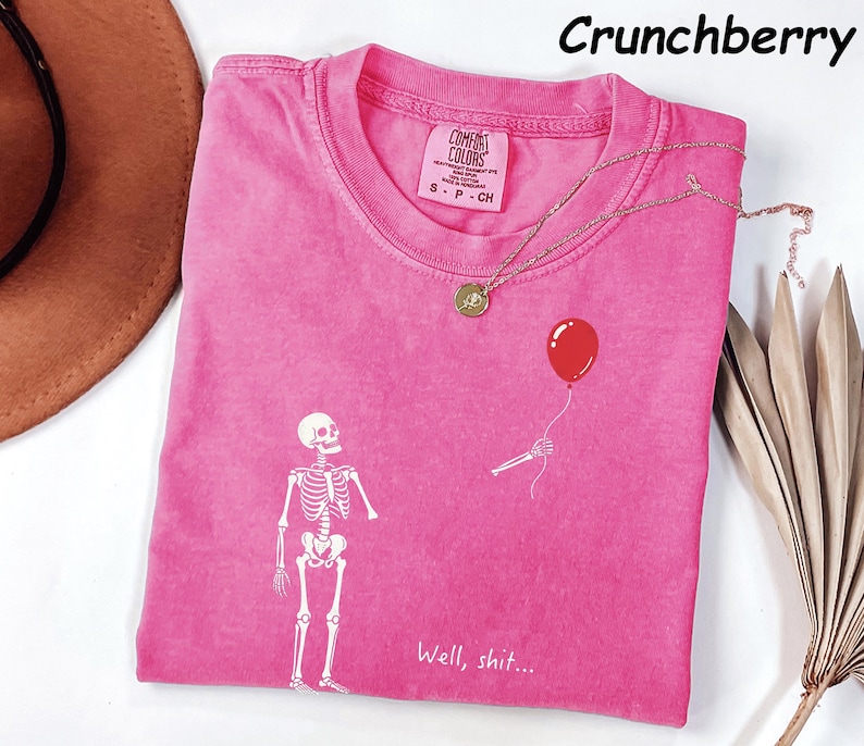 May include: A pink short-sleeved t-shirt featuring a white graphic of a skeleton looking up at a red balloon held by a skeletal hand. The text "Well, shit..." is printed in white below the skeleton. The shirt has a crew neck and a Comfort Colors label.