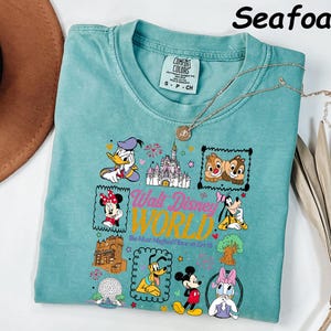 K&ouml;nnte beinhalten: T&uuml;rkisfarbenes T-Shirt mit einem farbenfrohen Grafikdesign von Disney-Figuren und dem Text "Walt Disney World". Das Design umfasst Donald Duck, Minnie Mouse, Chip und Chap, Goofy, Mickey Mouse und Daisy Duck. Das Shirt ist von der Marke Comfort Colors.